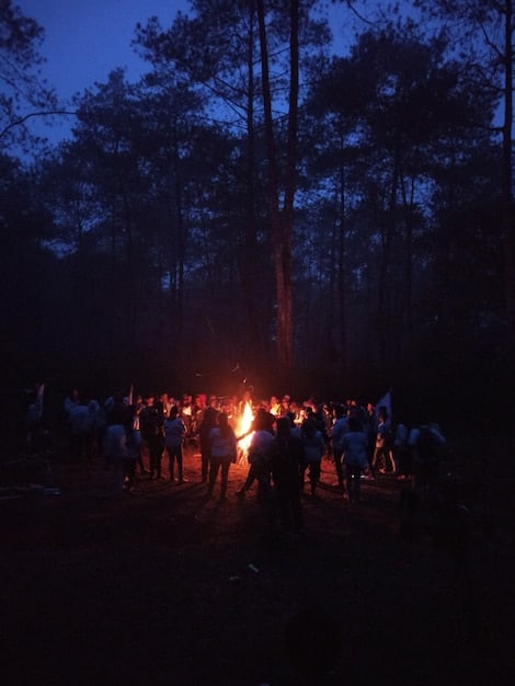 A group of survivors huddled around a campfire at night, their faces illuminated by the flickering flames, showing a mix of hope and weariness. One of them is playing a guitar, while others are listening intently. The background is dark and ominous.